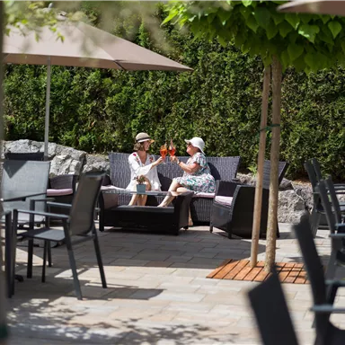 Two women are sitting comfortably in a garden area, enjoying drinks. Surrounded by sun umbrellas and trees, the atmosphere feels friendly and inviting.