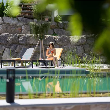 A woman is relaxing by the pool on a lounge chair. Surrounded by beautiful landscape design and clear water, she enjoys the tranquility.