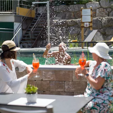 A relaxed atmosphere by the pool with two women holding drinks. In the background, a man is splashing in the water.