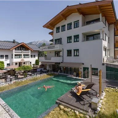 A modern hotel with a large terrace and a beautiful swimming pool. In the background, mountains and a clear blue sky can be seen.