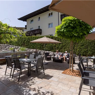 A cozy outdoor area with tables and chairs under sun umbrellas. In the background, green bushes and a building are visible.