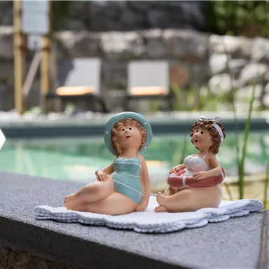 Two decorative figures of children at the pool's edge. One is sitting in a swimsuit, while the other is holding a flotation ring.