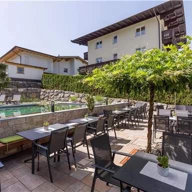 A lovely outdoor area with tables and chairs next to a pool. In the background, there are some buildings and trees.