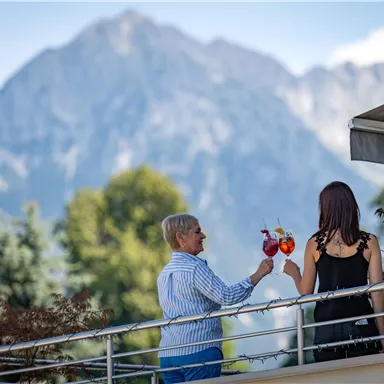 Two people on a balcony enjoy drinks with a view of the mountain landscape. The surroundings are green and the sky is clear.