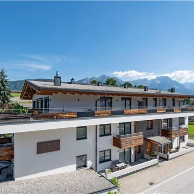 A modern building with wooden cladding in a picturesque landscape. In the background, mountains and a clear blue sky can be seen.