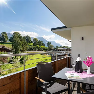 A beautiful balcony overlooking green meadows and mountains. A table with flowers and a bottle of wine invites you to relax.