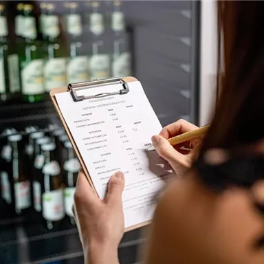 A person is holding a clipboard and writing on a list. In the background, shelves with bottles are visible.