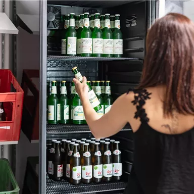 A person is standing in front of a refrigerator with various beer bottles. Empty crates are visible in the background.