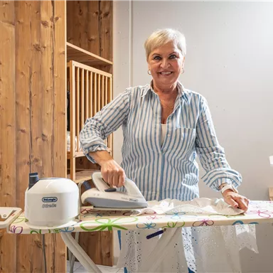A friendly woman is ironing in a bright room. In the background, a laundry basket and a washing machine can be seen.