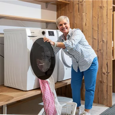 An older woman stands smiling next to a washing machine and pulls laundry out. The room is modern and bright, with wooden shelves in the background.