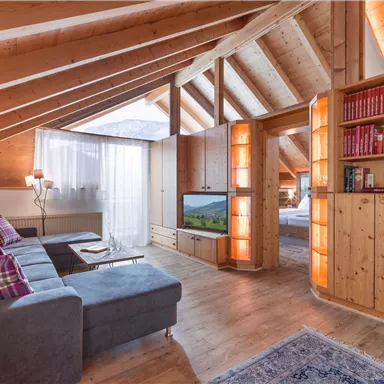 A cozy loft with wooden paneling and large windows. The living area is bright and modernly furnished, with a television and a bookshelf.