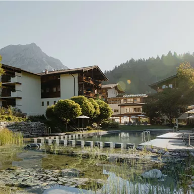 A picturesque hotel in the mountains, surrounded by trees and shrubs. In the foreground is a tranquil pond with modern loungers and umbrellas.
