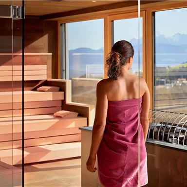 A woman in a pink towel stands in front of large windows in a modern wellness area. In the background, mountains and a serene landscape are visible.