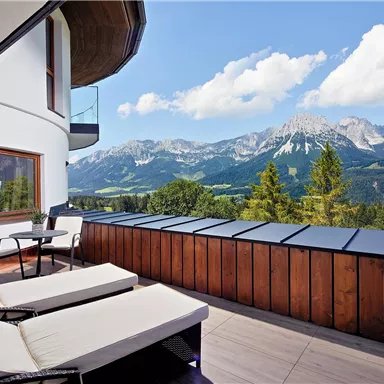 An elegant balcony with loungers and a table. Impressive mountains and a clear blue sky can be seen in the background.