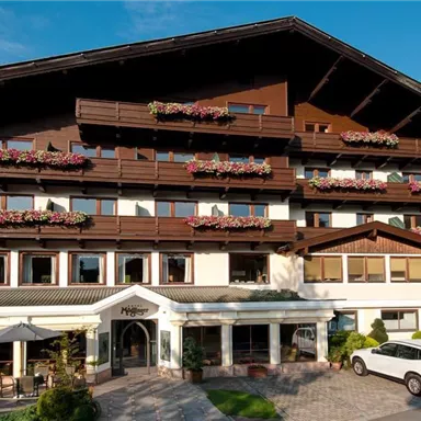 A charming hotel building with balconies and floral decorations. The sunny surroundings and the mountain landscape give the place an inviting atmosphere.