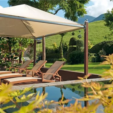 A cozy garden with loungers under a sunshade. In the background, green hills and well-maintained plants can be seen.