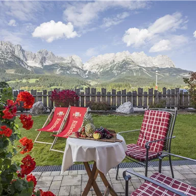 A cozy terrace with a view of majestic mountains. In the foreground, there are blooming flowers and a table with fresh fruits.