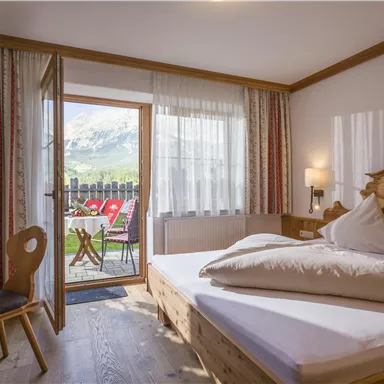 A cozy bedroom with a double bed and wooden furniture. A balcony with a view of nature leads from the door.