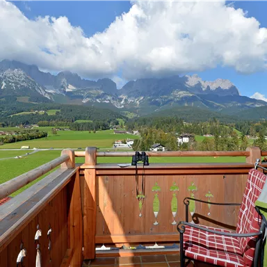 A balcony with a view of majestic mountains and a green landscape. The clouds are light and the sky is blue.