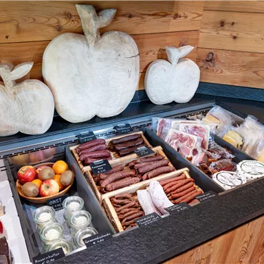 Eine Auswahl an Wurstwaren und Snacks liegt auf einem Holzregal. Im Hintergrund sind dekorative Holzäpfel zu sehen.