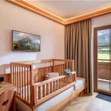 Ein helles Zimmer mit einem Kinderbett aus Holz und großem Fenster. Der Raum bietet einen Blick auf die Berge und einen Balkon.