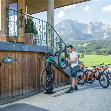 Ein Mann repariert ein Fahrrad vor einem Holzgebäude in einer malerischen Berglandschaft. Im Hintergrund sind grüne Wiesen und hohe Berge sichtbar.