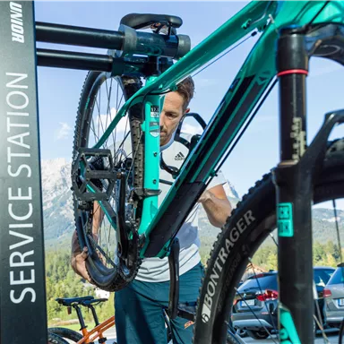 Eine Person arbeitet an einem Fahrrad in einer Service-Station. Im Hintergrund sind Berge und Autos zu sehen.