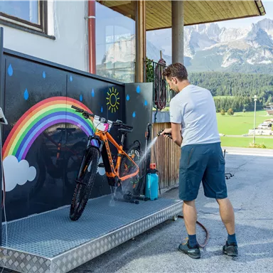 Ein Mann wäscht ein Fahrrad an einer Waschstation mit einem Regenbogen Design. Im Hintergrund sind Berge und eine grüne Landschaft zu sehen.