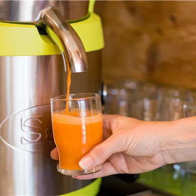 Ein Getränkespender mit frischem, orangefarbenem Saft wird in ein Glas gefüllt. Eine Hand hält das Glas, während der Saft ausgeschenkt wird.