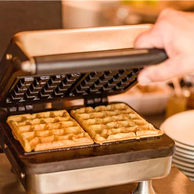 Ein Waffeleisen mit frischen, goldbraunen Waffeln wird geöffnet. Im Hintergrund sind Geschirr und Zutaten sichtbar.