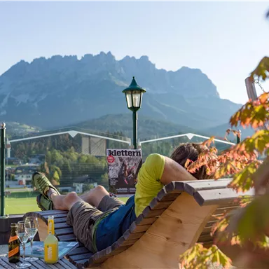 Ein entspannter Mann liegt auf einer Liege und liest eine Zeitschrift. Im Hintergrund sind beeindruckende Berge und eine klare, sonnige Landschaft zu sehen.