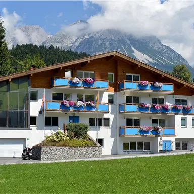 A modern building with balconies and blooming plants. In the background, tall mountains and a blue sky are visible.