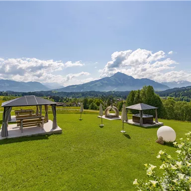 Eine malerische Landschaft mit grünem Gras und Blick auf die Berge. Im Vordergrund befinden sich moderne Pavillons und Gartenmöbel.