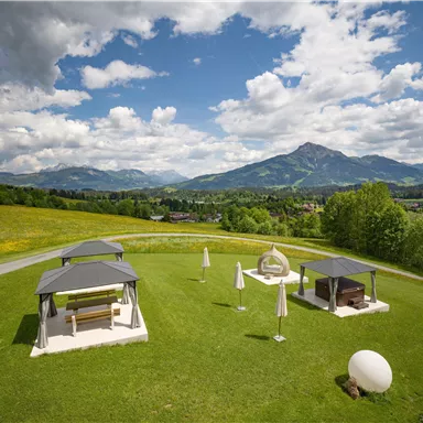 Eine malerische Landschaft mit grünen Wiesen und Bergen im Hintergrund. Zwei Pavillons stehen auf einer Wiese unter einem klaren, blauen Himmel.