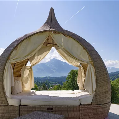 Ein gemütliches Daybed im Freien mit einem Blick auf die Berge und den blauen Himmel. Die Umgebung ist grün und einladend.