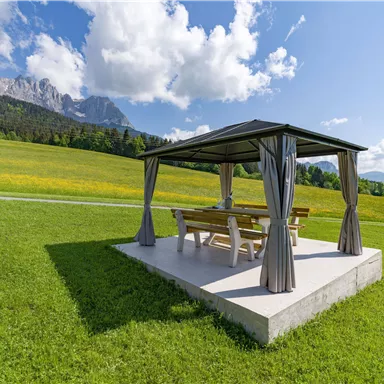 Ein Pavillon auf einer Wiese mit einer Aussicht auf Berge. Die Umgebung ist grün und sonnig.