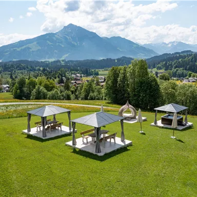 Eine grüne Wiese mit mehreren Pavillons und Blick auf die Berge. Der Himmel ist blau und teilweise bewölkt.