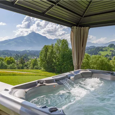 Ein Whirlpool unter einem Pavillon mit Blick auf die Berge. Die Umgebung ist grün und bietet eine ruhige, entspannende Atmosphäre.