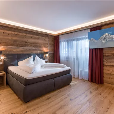 Ein modernes Schlafzimmer mit einem großen Bett und Holzverkleidung. Ein Fernseher zeigt ein Bild von Bergen, und es gibt Fenster mit Vorhängen.