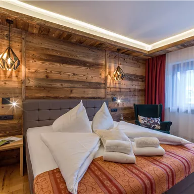 A cozy bedroom with wooden walls and modern lighting accents. The bed is adorned with white pillows and a red blanket, next to the bed stands a small table.