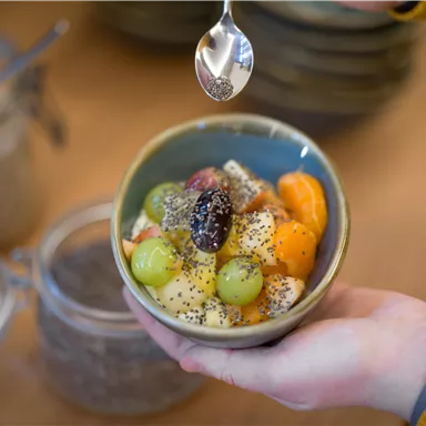 A bowl of fresh fruit, including grapes, mandarins, and bananas. Someone is pouring chia seeds over it.