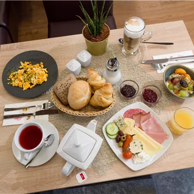 A hearty breakfast buffet with scrambled eggs, rolls, cold cuts, and fresh fruit. Accompanied by tea, jam, and juice served on a wooden table.