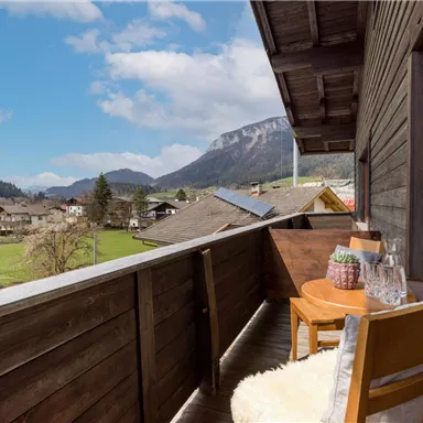 A balcony with a wooden railing overlooking a rural landscape. In the foreground, there are two chairs and a table with a plant.