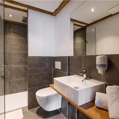 Ein modernes Badezimmer mit einer klaren Dusche und einer schlichten Waschschüssel. Es sind Handtücher und Seifenspender vorhanden, sowie elegante Holzdetails.