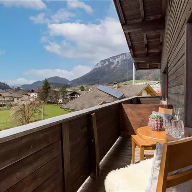 Ein Balkon mit Holzgeländer und Blick auf eine ländliche Landschaft. Im Vordergrund stehen zwei Stühle und ein Tisch mit einer Pflanze.