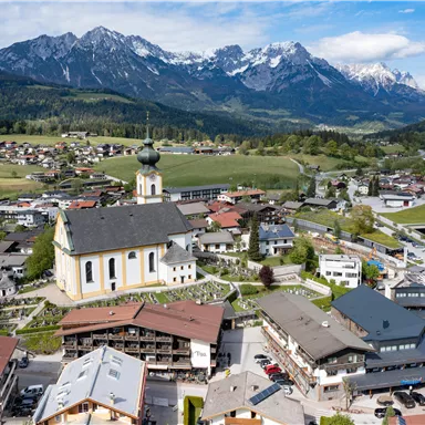 A picturesque town with a beautiful church in the foreground and majestic mountains in the background. The landscape is green and peaceful, ideal for outdoor activities.