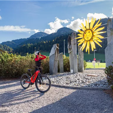 A cyclist stands next to an artistic sun sculpture and natural stone monuments. In the background, green meadows and mountains can be seen.