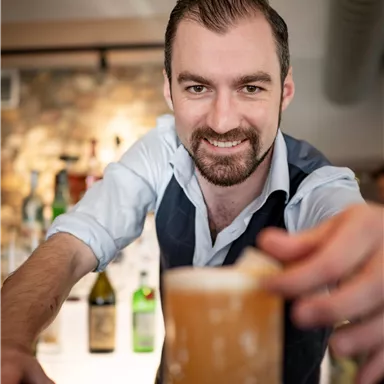 A bartender presents a drink in a bar. In the background, bottles and a cozy atmosphere can be seen.