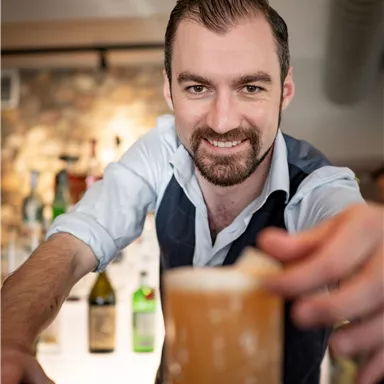 Ein Barkeeper präsentiert ein Getränk in einer Bar. Im Hintergrund sind Flaschen und eine gemütliche Atmosphäre zu sehen.