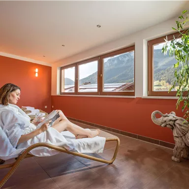 Eine Frau entspannt in einem Liegestuhl und liest ein Buch in einem gemütlichen Raum. Große Fenster bieten einen Blick auf die Berge im Hintergrund.